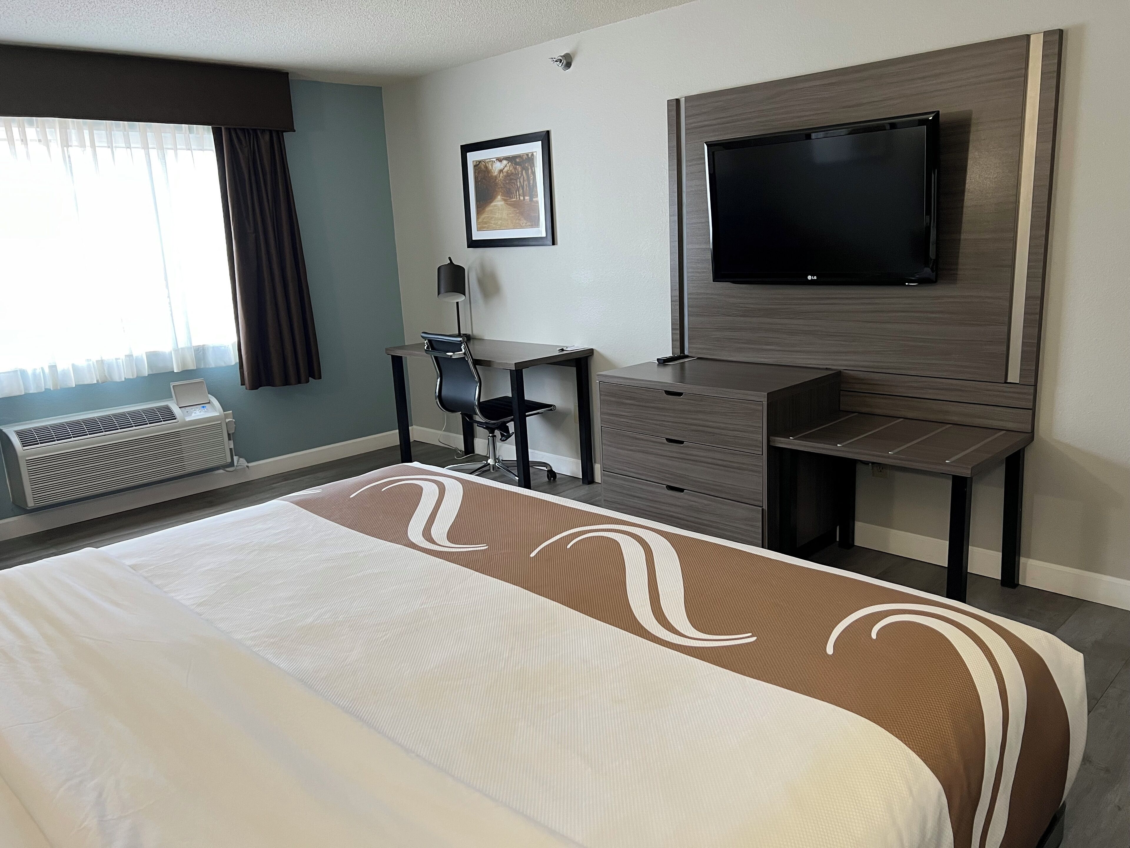 A hotel room with a large bed, a desk with a chair, a flat-screen TV mounted on the wall, and a window with curtains.