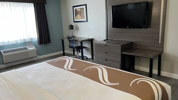 A hotel room with a large bed, a desk with a chair, a flat-screen TV mounted on the wall, and a window with curtains.