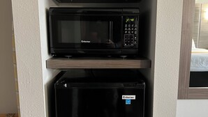 A compact kitchen area with a black microwave and refrigerator, a shelf with kitchen items, and a wooden door frame.