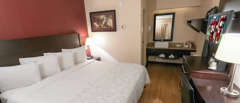 A hotel room with a bed, a desk with a lamp, a mirror, a bathroom with a sink, and a television.
