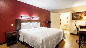 Hotellihuone, jossa on sänky, yöpöytä, työpöytä, tuoli ja ovi, joka johtaa kylpyhuoneeseen.