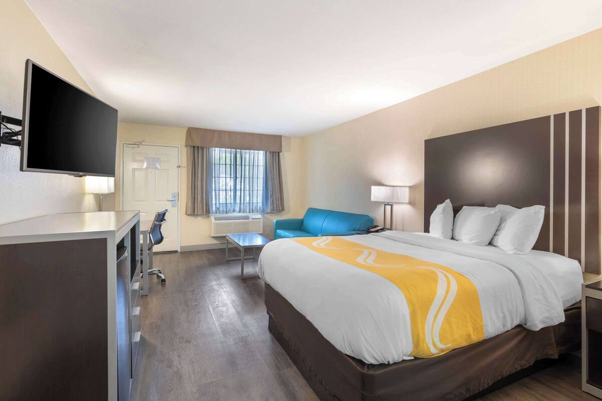 Quarto de hotel com uma cama grande, escrivaninha, televisão de tela plana, poltrona azul e janela com cortinas.