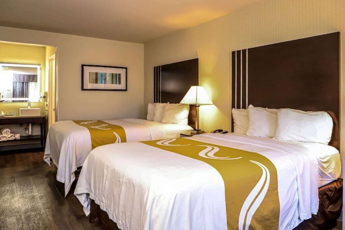 Quarto de hotel com duas camas, uma mesinha de cabeceira, uma luminária, um quadro na parede e um banheiro visível por uma porta aberta.