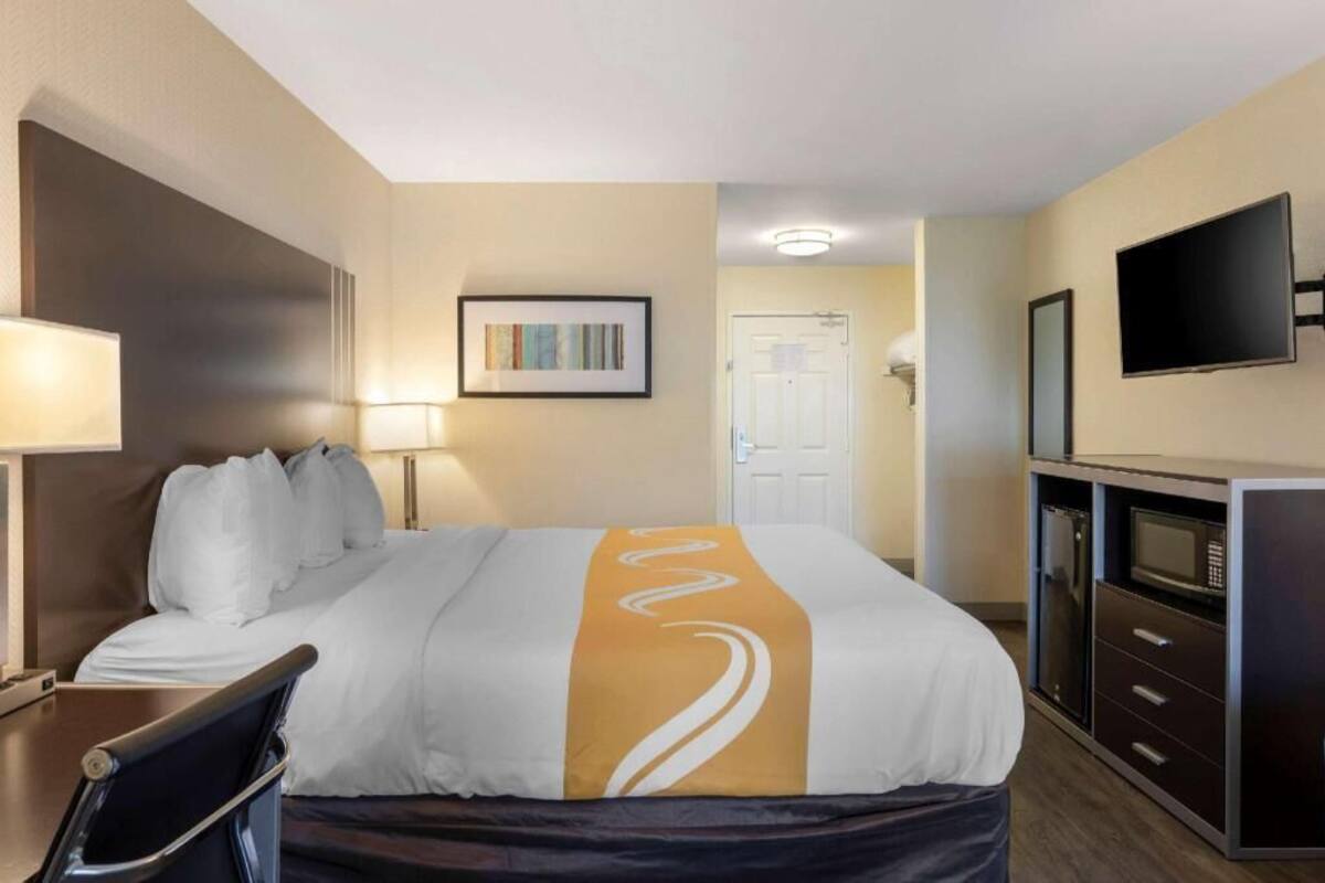 Quarto de hotel com uma cama, escrivaninha, cadeira, televisão, micro-ondas e porta.