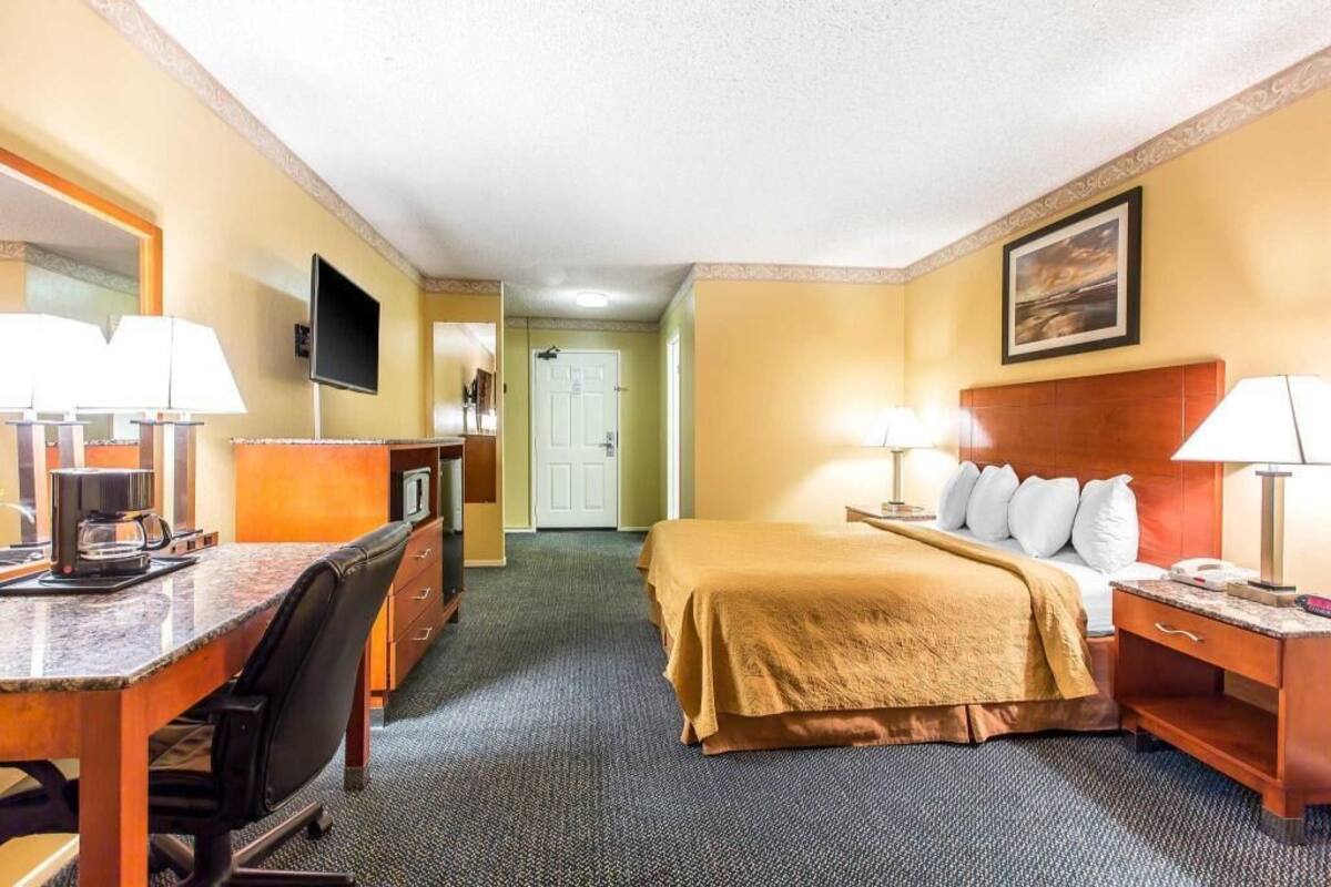 Quarto de hotel com cama, escrivaninha, cadeira, televisão e uma porta.