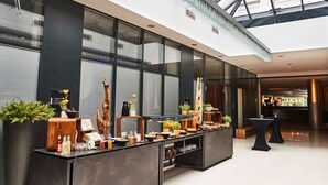 A modern hotel lobby with a glass ceiling, a reception desk with a coffee station, and a seating area with tables.