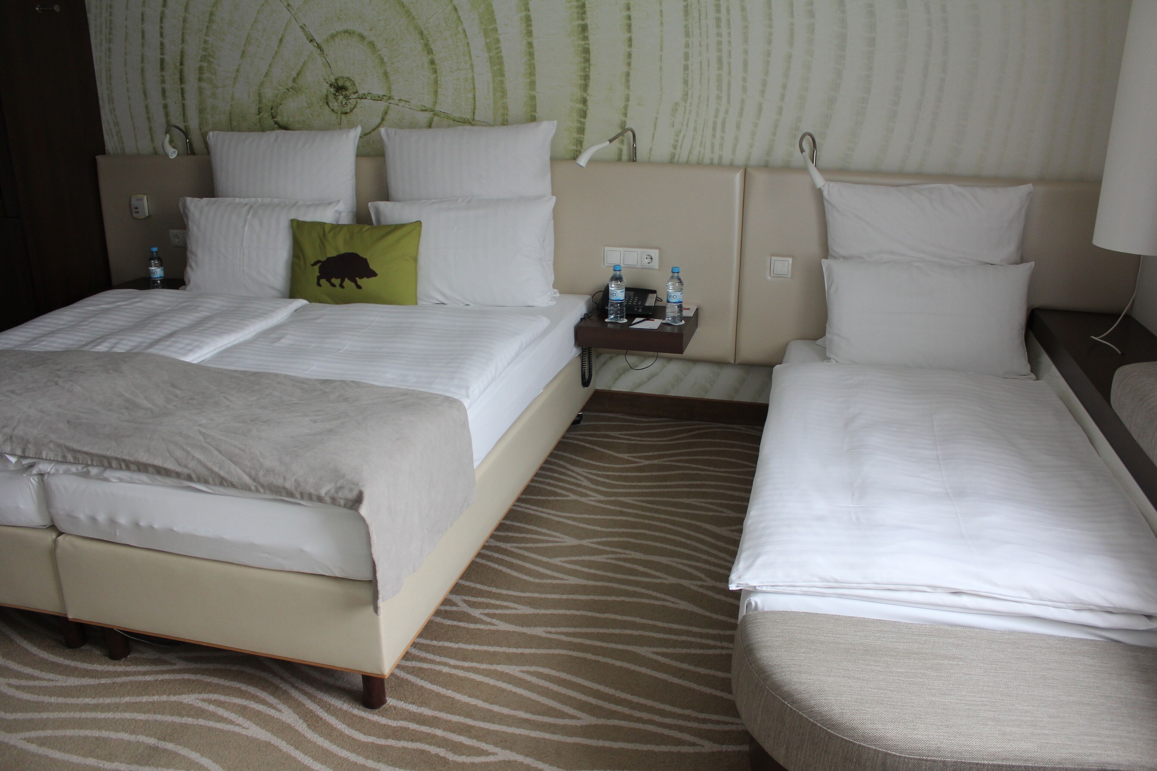 Ein Hotelzimmer mit einem großen Bett, einem kleineren Bett, einem Nachttisch mit einer Lampe und einer Wand mit einem grünen abstrakten Muster.