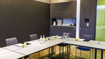 A conference room with a long table, chairs, a shelf with cups and a kettle, and a projector screen.