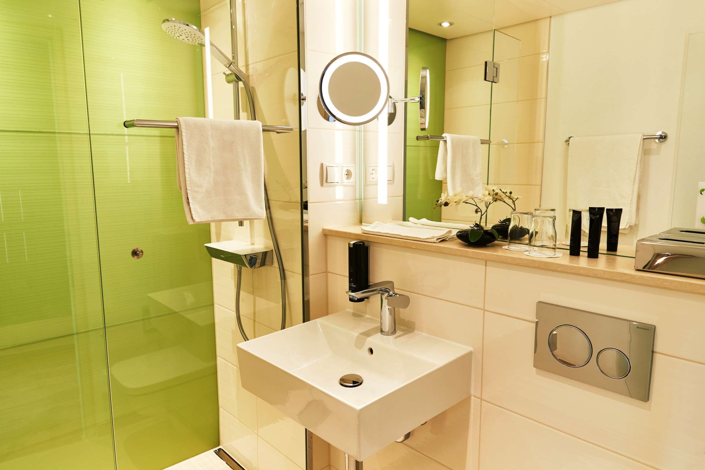Un baño moderno con un lavamanos blanco, cabina de ducha con mampara de vidrio y pared revestida de azulejos verdes.