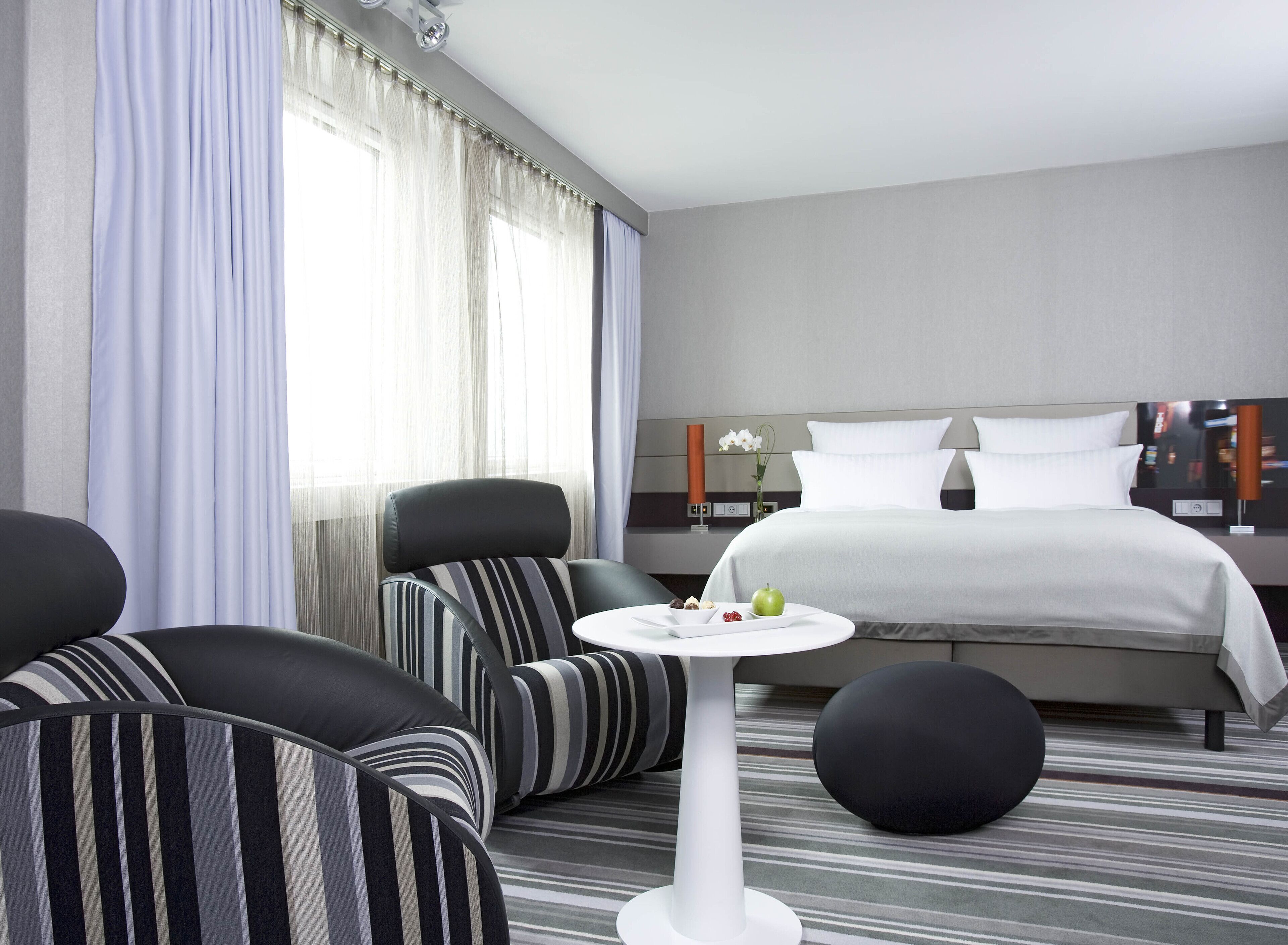 Una habitación de hotel con cama, dos sillones, una mesita redonda con una bandeja de frutas y un ventanal con cortinas traslúcidas.