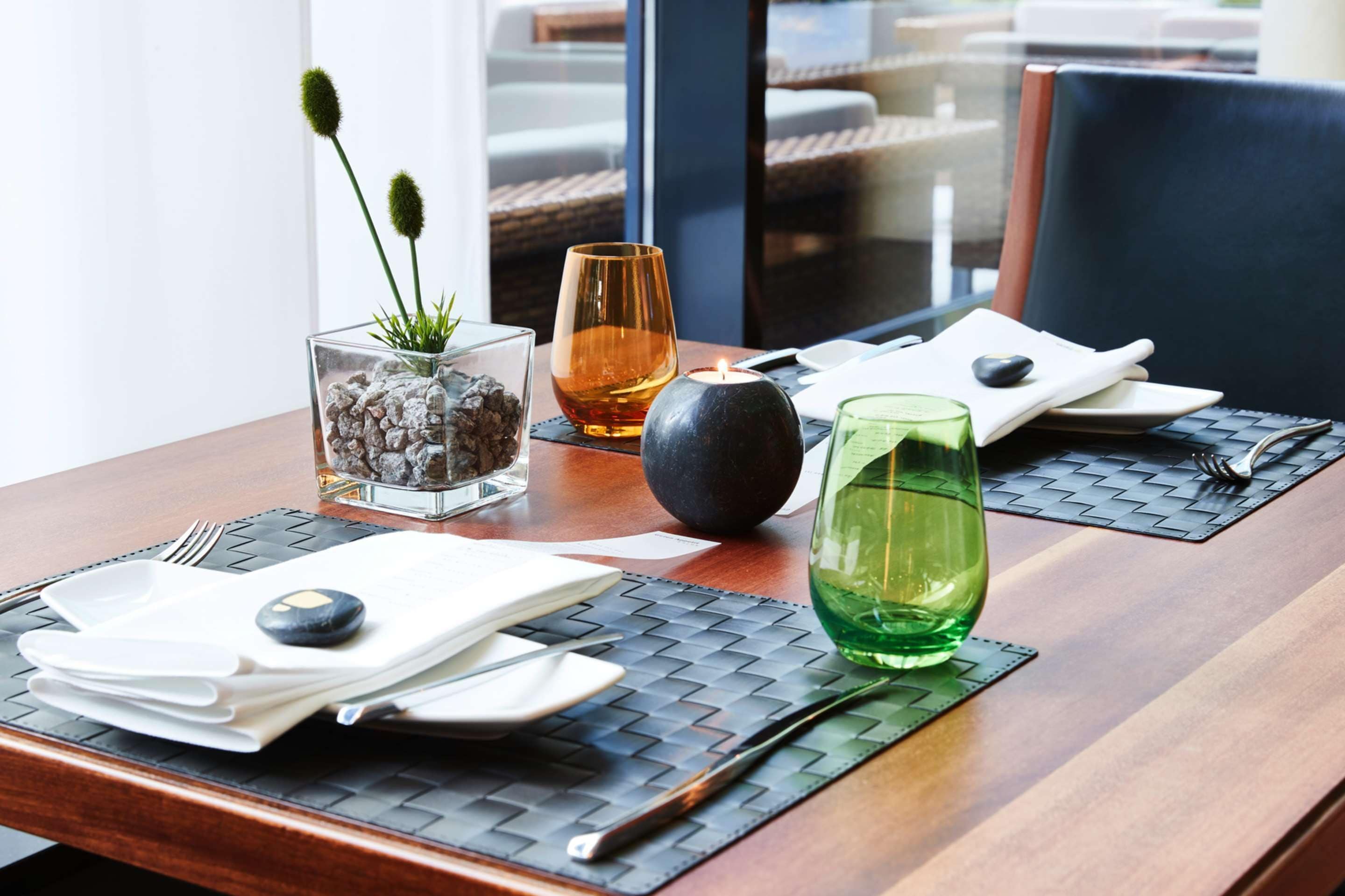 Ein Esstisch mit Kerze, einem Glas Wasser, einem Glas bernsteinfarbener Flüssigkeit und einer kleinen Pflanze in einem Glasgefäß.