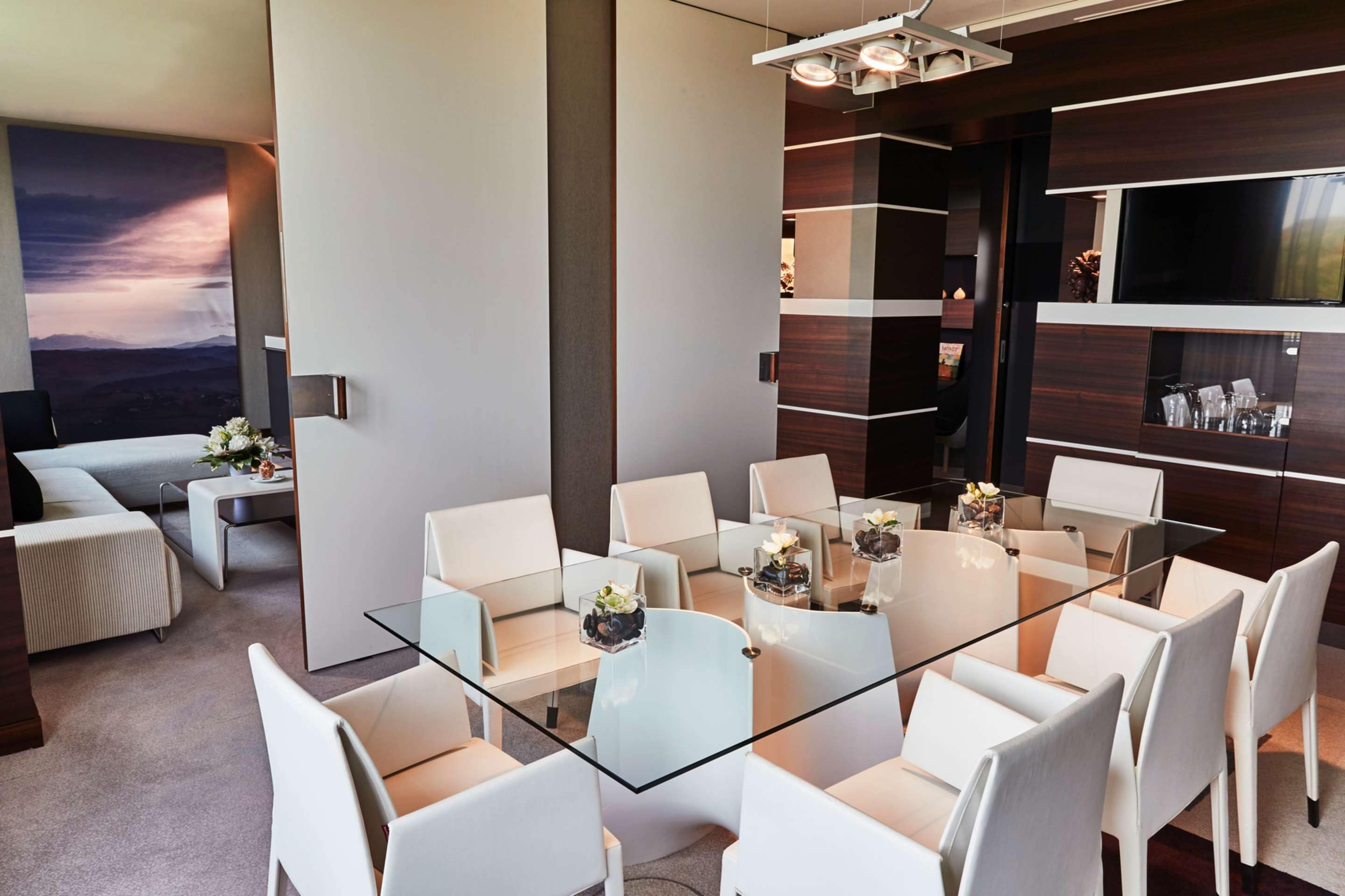 A modern conference room with a glass table, white chairs, and a large TV on the wall.