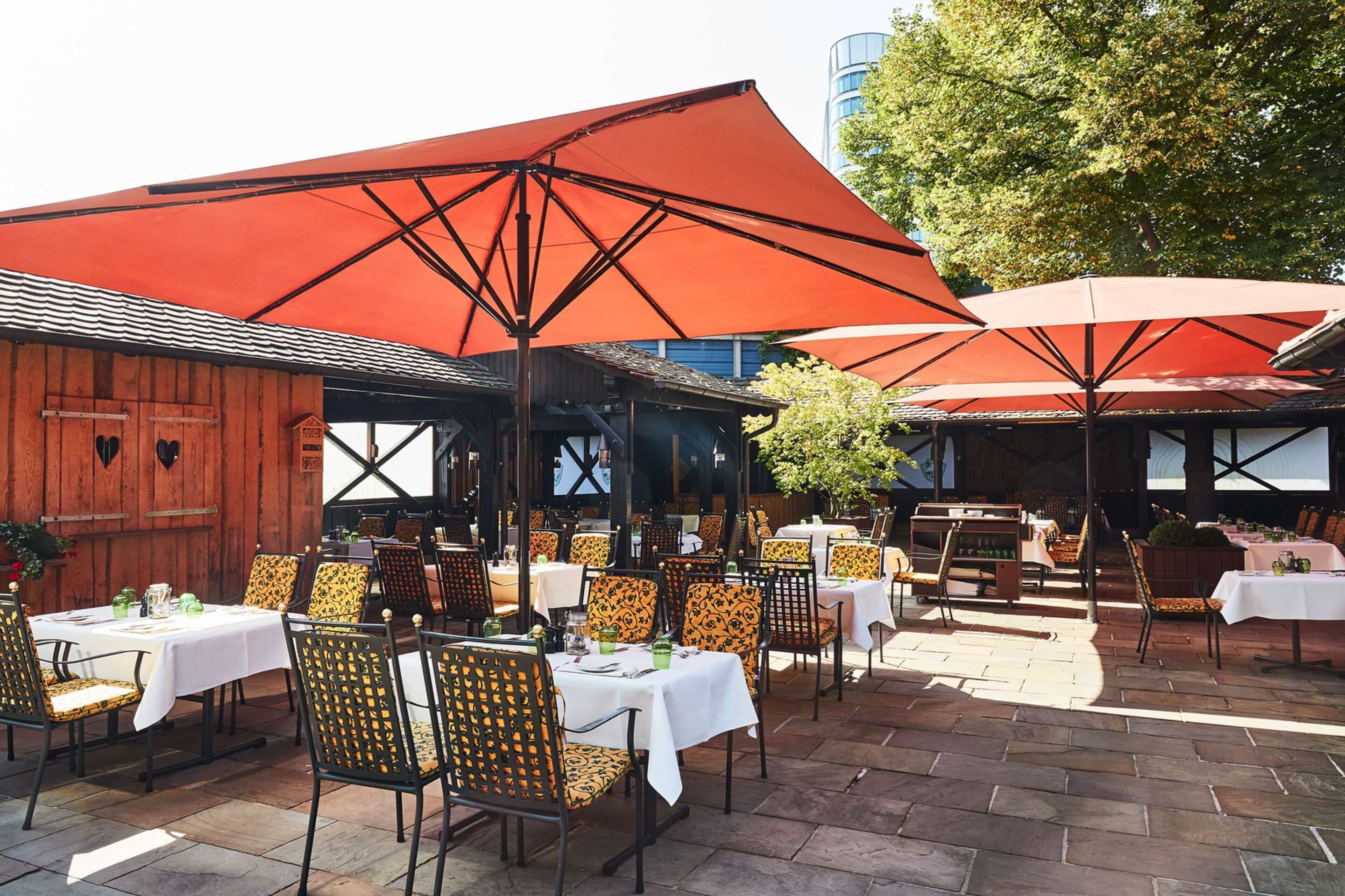 Un espace extérieur aménagé pour les repas, avec des tables et des chaises, protégé par de grands parasols rouges, et un bâtiment en bois en arrière-plan.