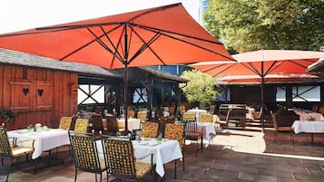Un espace extérieur aménagé pour les repas, avec des tables et des chaises, protégé par de grands parasols rouges, et un bâtiment en bois en arrière-plan.