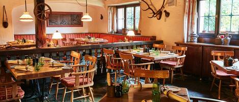 A rustic dining area with wooden tables and chairs, a large wooden beam ceiling, and a window with curtains.