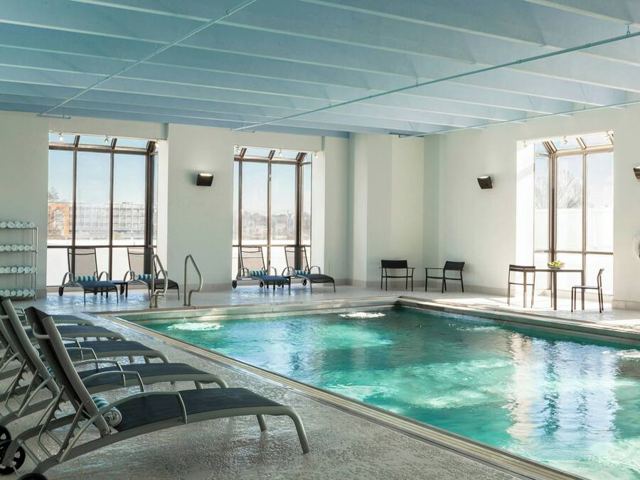 An indoor swimming pool with clear blue water, sunlit by large windows, and surrounded by lounge chairs and tables.