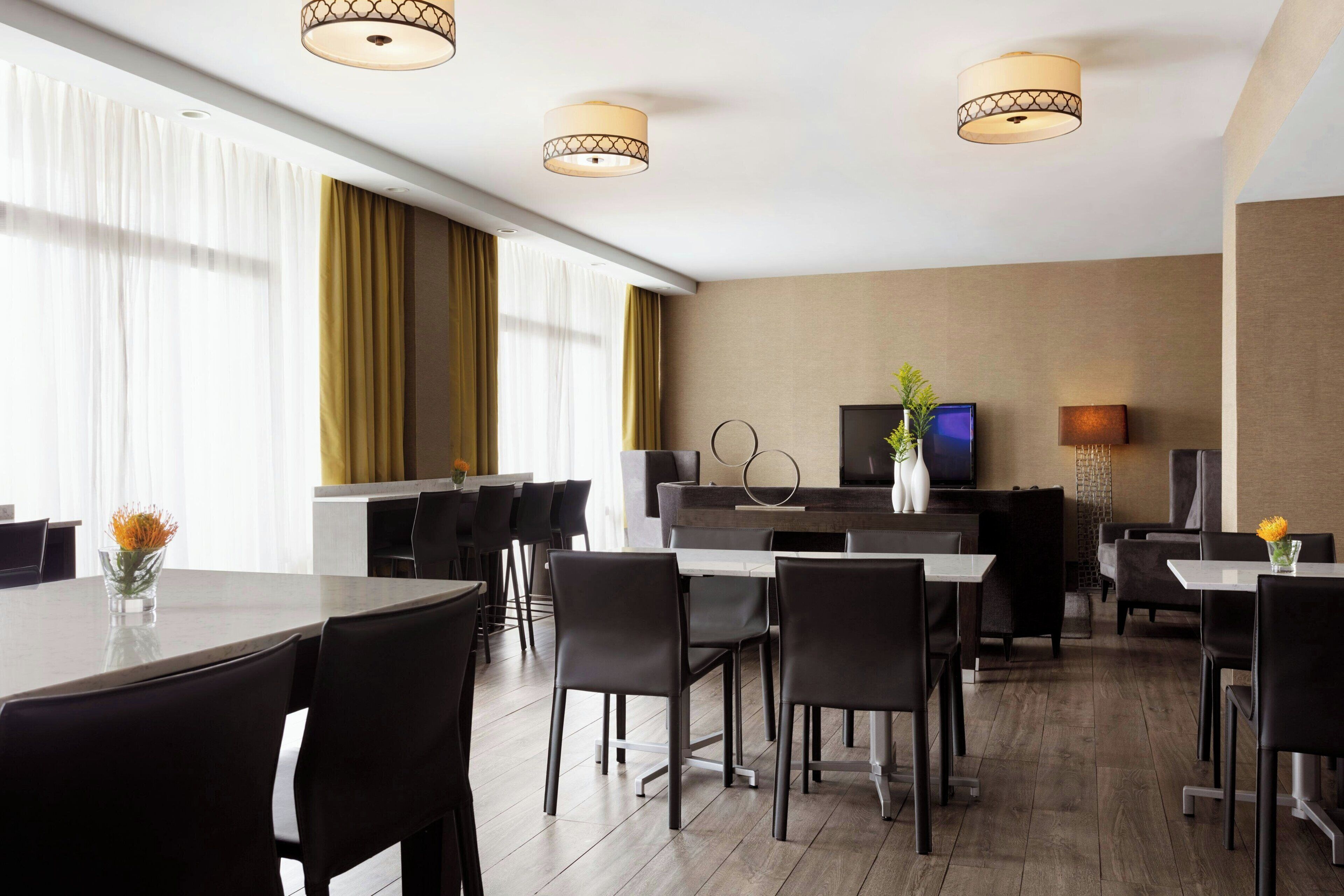 A modern hotel dining area with a long table, several chairs, and a TV screen.