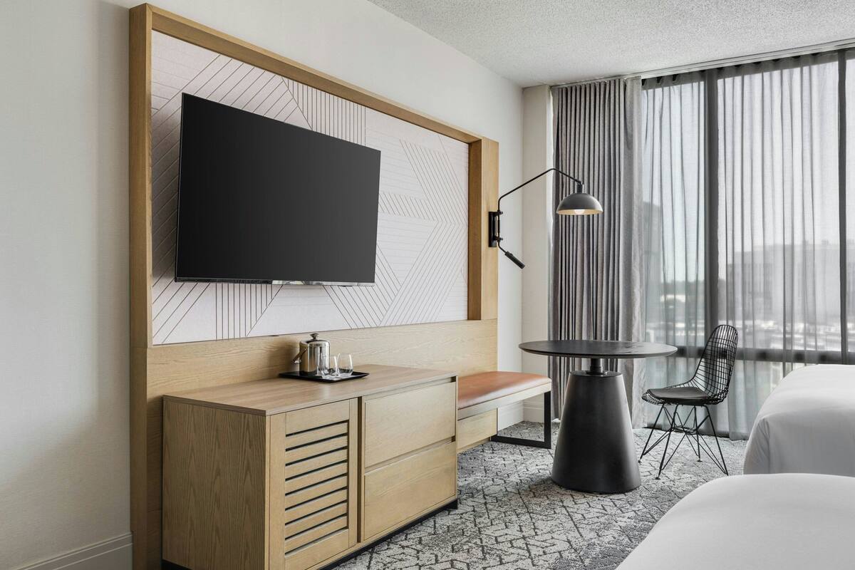 Una habitación de hotel moderna con un televisor de pantalla plana grande montado en un panel de madera, una mesita redonda con una lámpara, una silla y una cama con ropa de cama blanca.