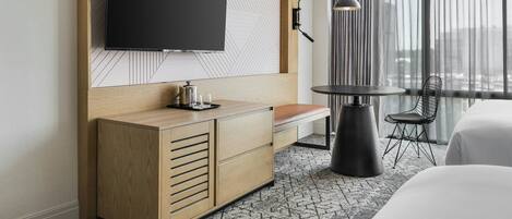 A modern hotel room with a large flat-screen TV mounted on a wooden panel, a small round table with a lamp, a chair, and a bed with white bedding.