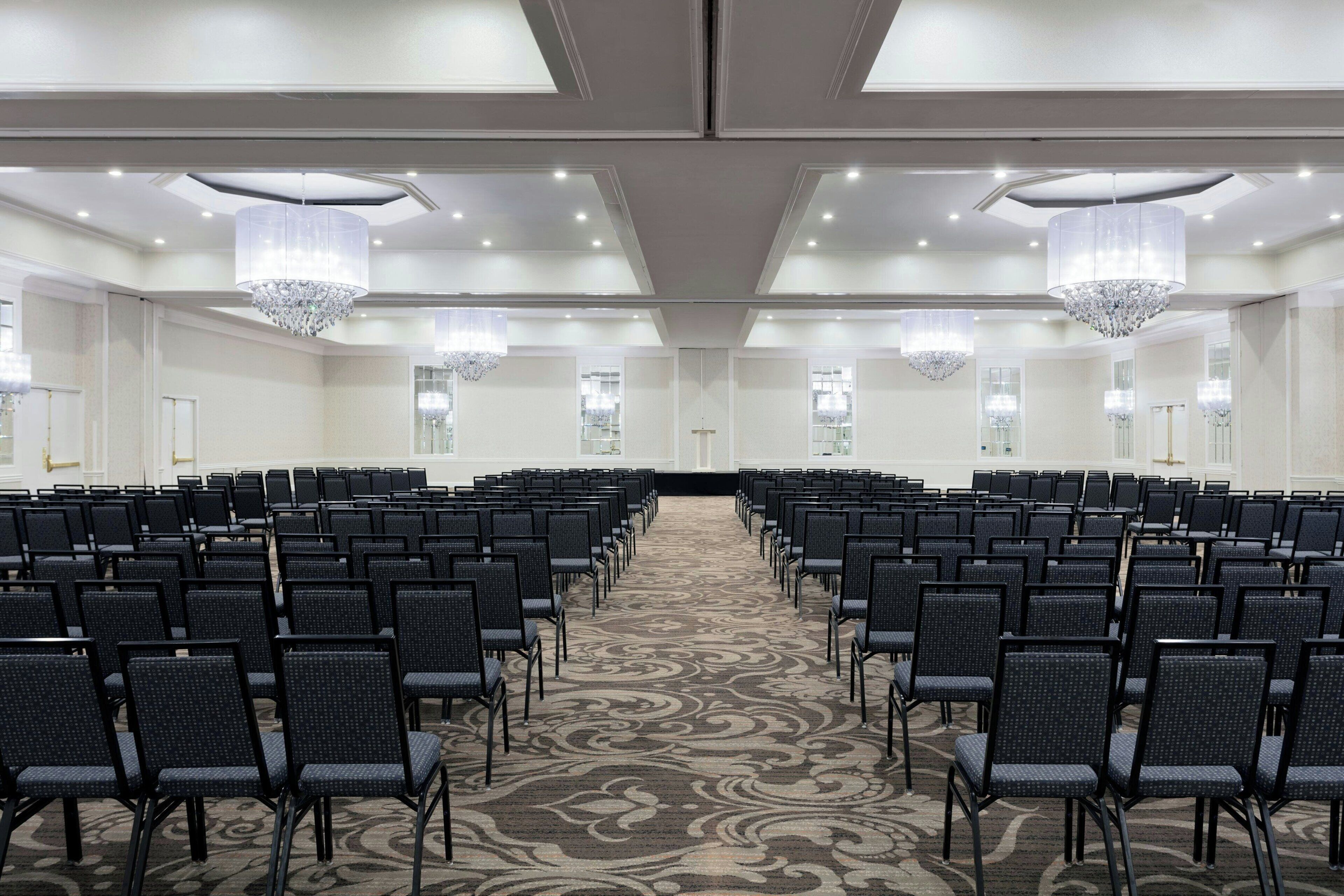 A conference room with rows of chairs, a patterned carpet, chandeliers, and large windows.