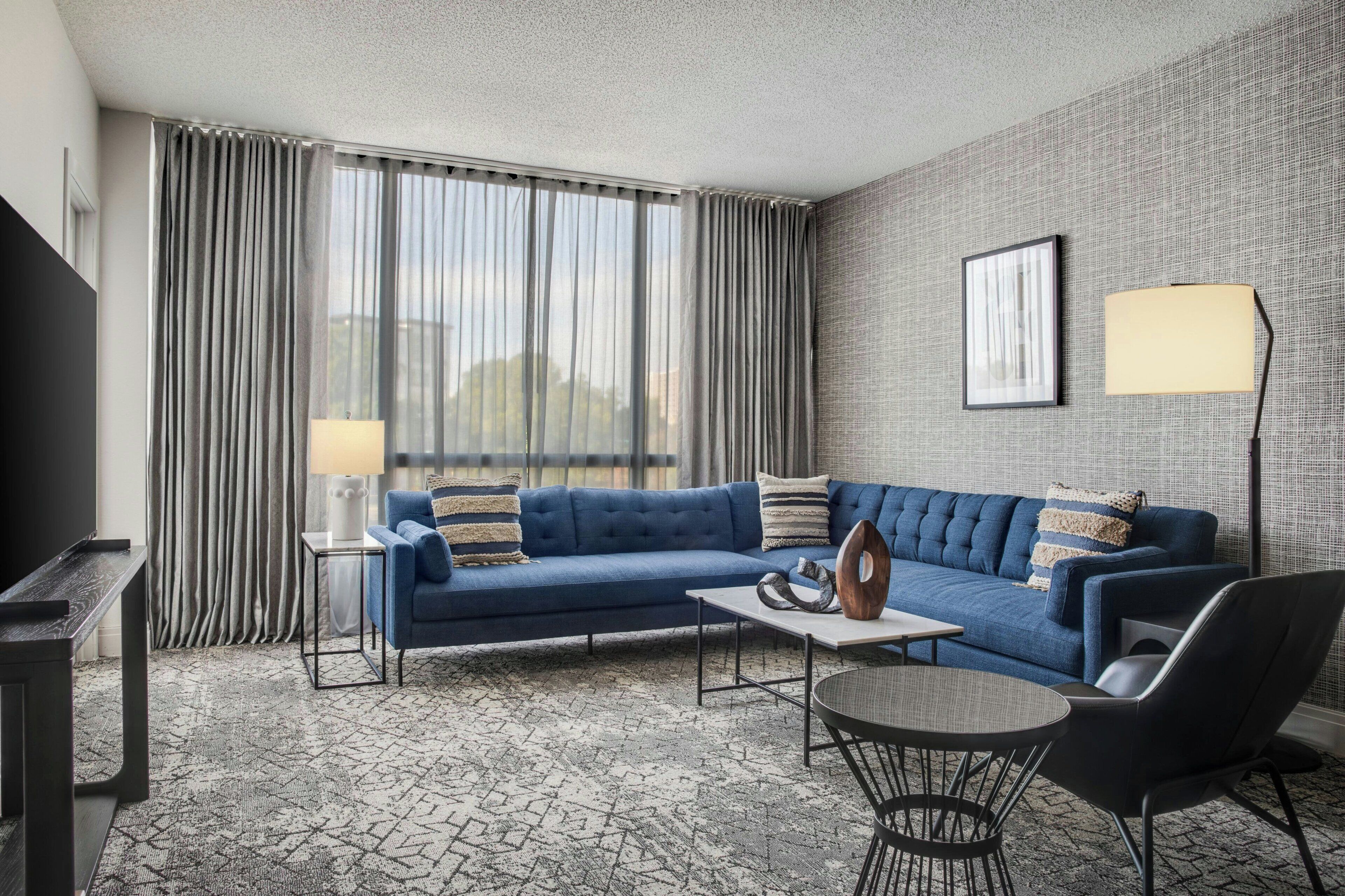 Et moderne hotelværelse med en blå hjørnesofa, et lille rundt bord, en sort lænestol, et fjernsyn og et stort vindue med gardiner.