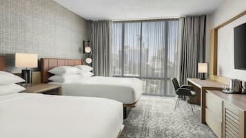 Habitación de hotel con dos camas, un escritorio con lámpara, una silla, un televisor y un ventanal con cortinas.