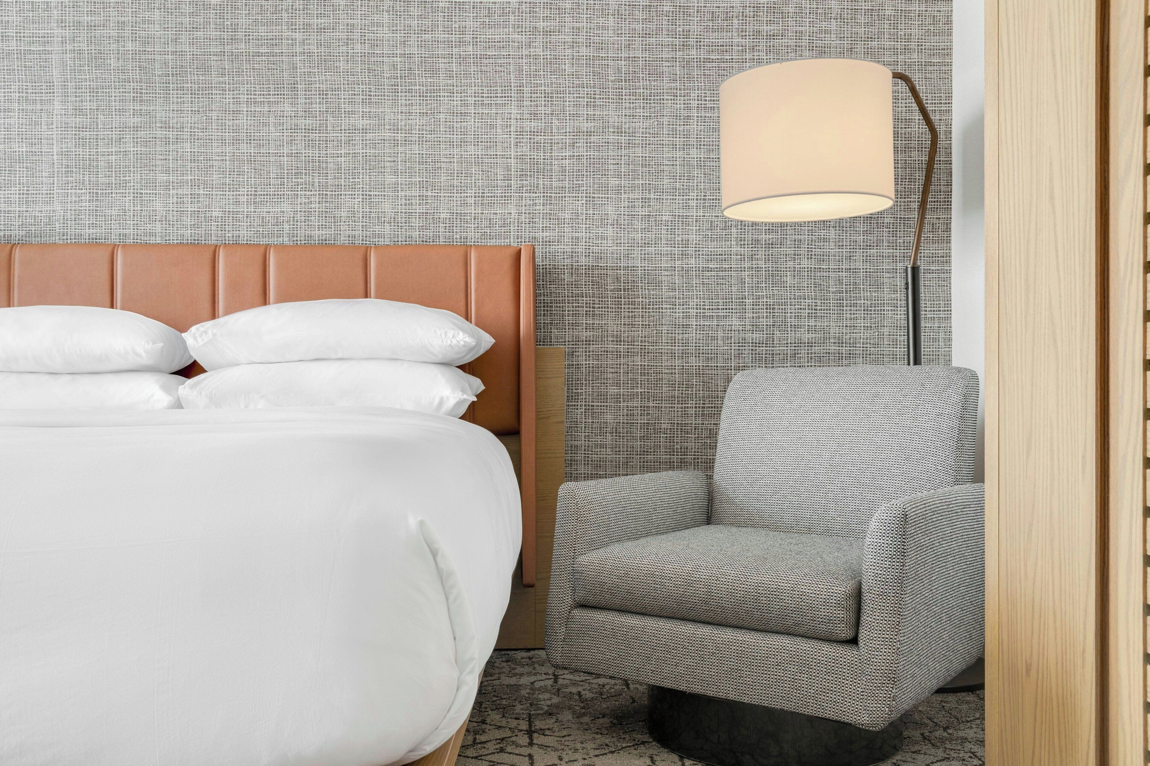 Una cama bien hecha con sábanas blancas, un sillón tapizado en gris y una lámpara de pie con pantalla color beige.