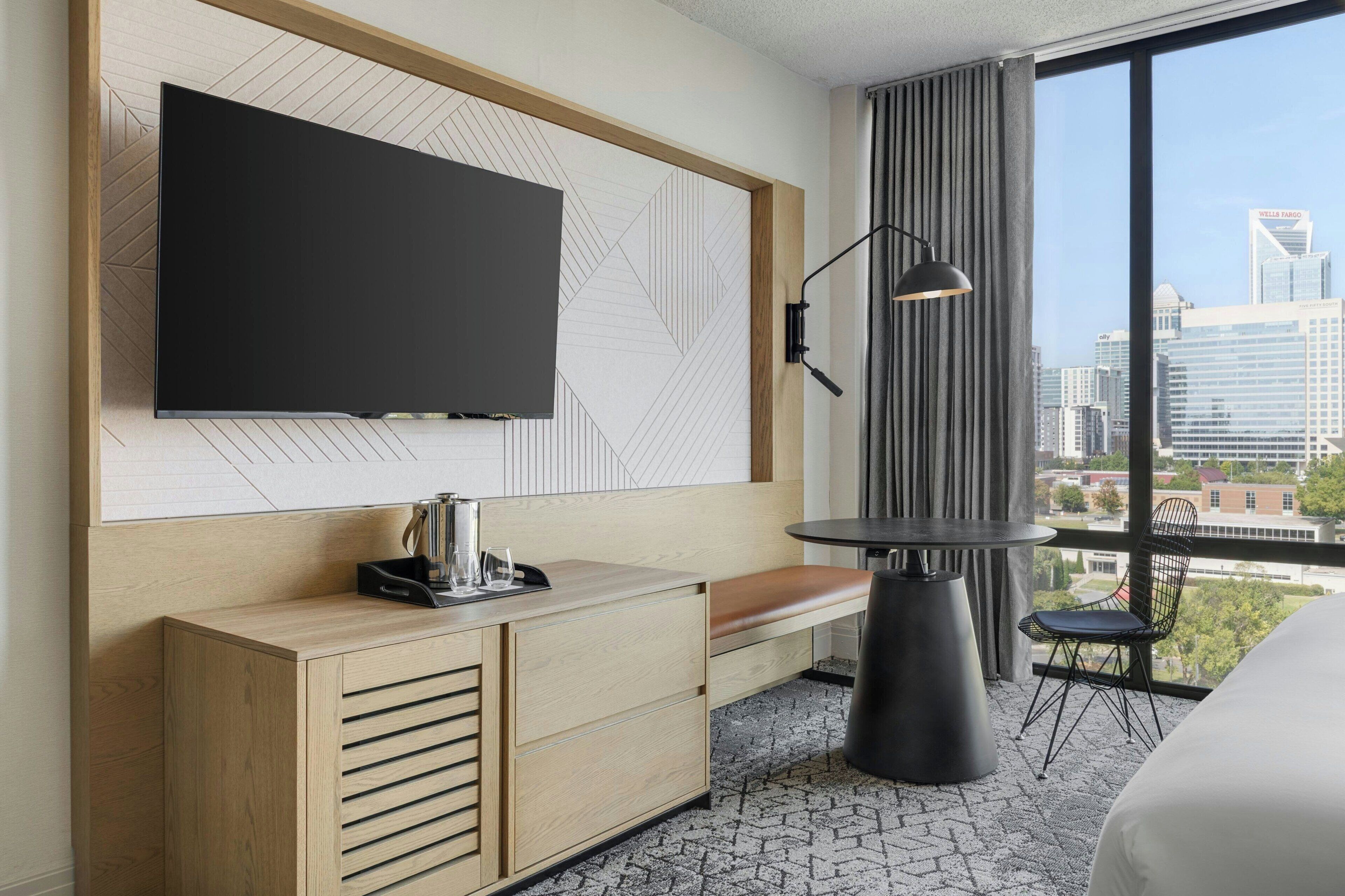 Ein Hotelzimmer mit einem großen Flachbildfernseher an der Wand, einer Holzkassette mit Schubladen, einer Bank, einem kleinen runden Tisch und Blick auf die Stadtlandschaft durch große Fenster.