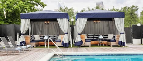 A poolside area with two covered seating areas, each with a canopy and curtains, and a pool with a wooden deck and lounge chairs.