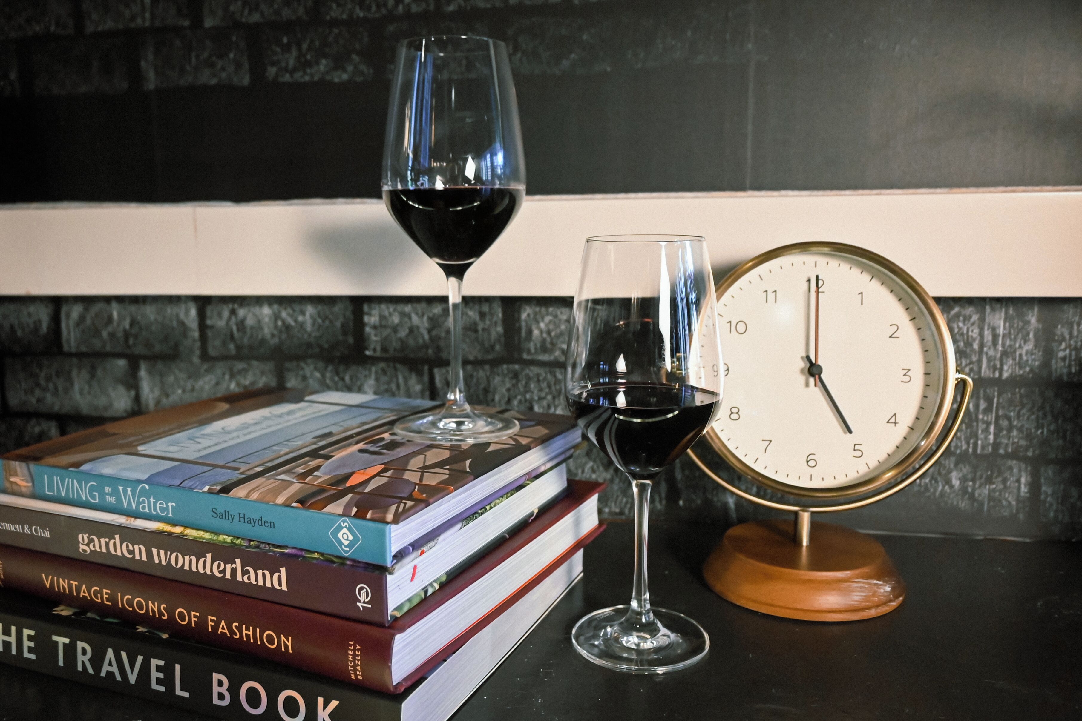 A stack of books, two wine glasses with red wine, and a vintage clock on a table.