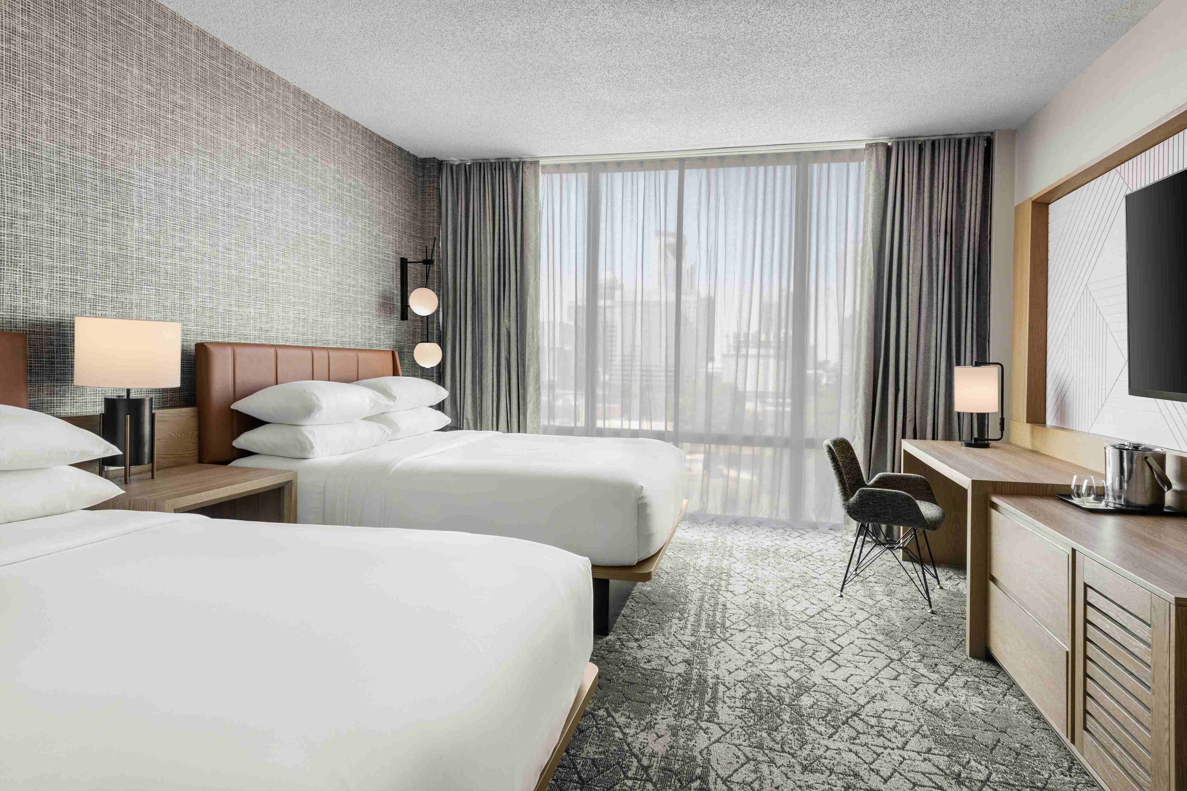 Ein Hotelzimmer mit zwei Betten, einem Schreibtisch, einem Sessel und einem Fernseher, mit Blick auf die Stadt.
