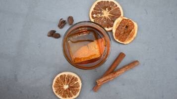 Un vaso de líquido ámbar con palitos de canela y rodajas de naranja deshidratada sobre una superficie gris.