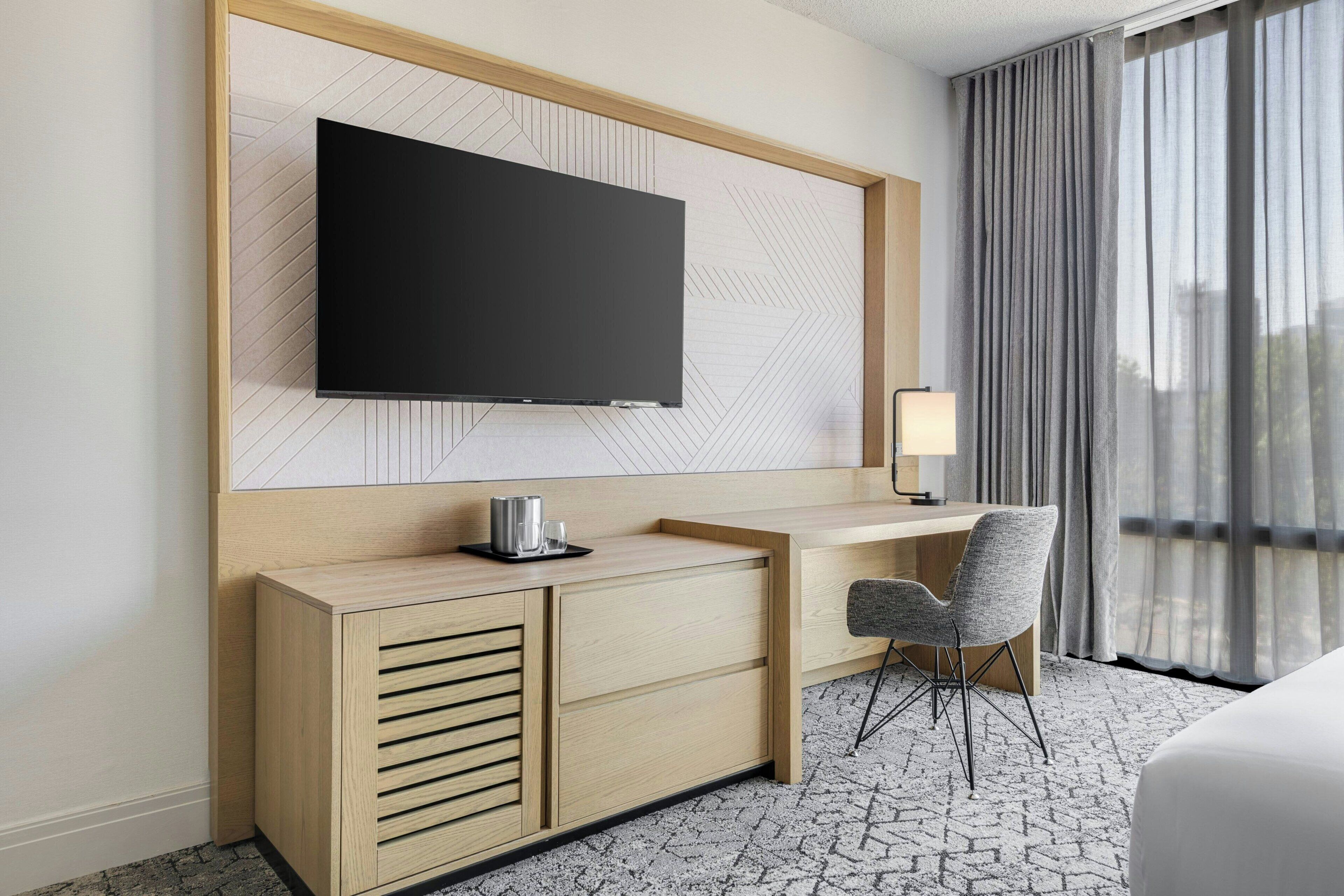 Ein Hotelzimmer mit Flachbildfernseher an einer Holzvertäfelung, einem Schreibtisch mit Stuhl, einem Nachttisch und einer Lampe.
