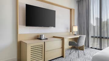 Ein Hotelzimmer mit Flachbildfernseher an einer Holzvertäfelung, einem Schreibtisch mit Stuhl, einem Nachttisch und einer Lampe.