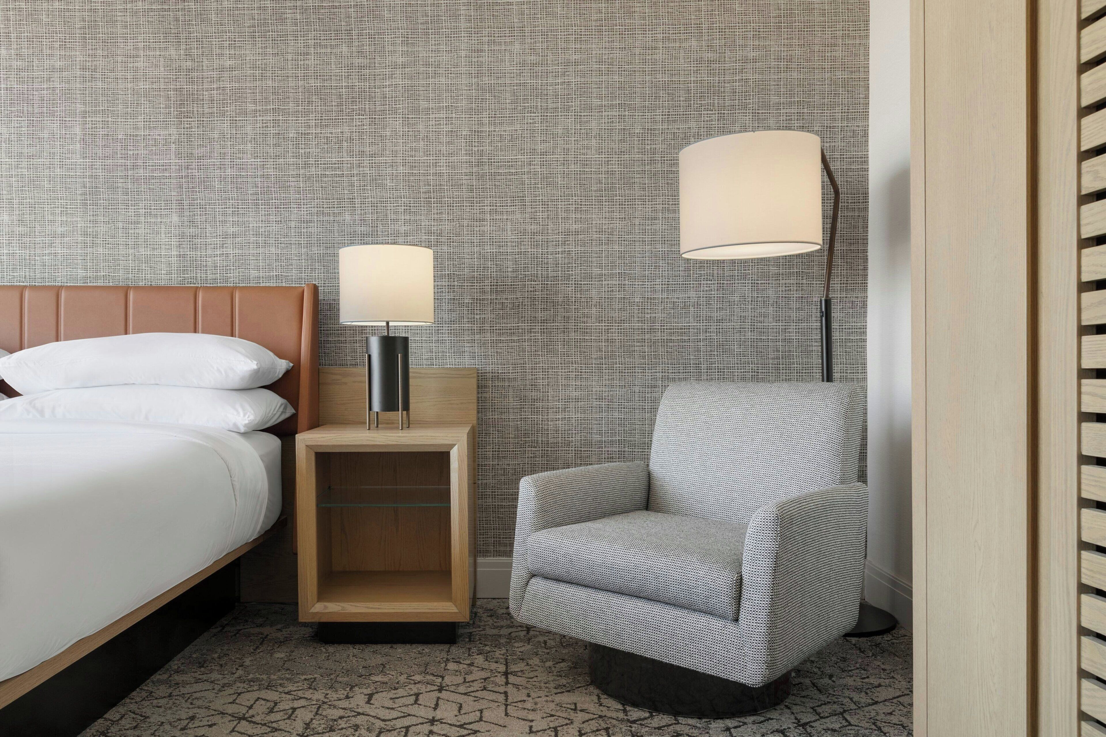 A hotel room with a bed, a nightstand with a lamp, a chair, and a textured wall.