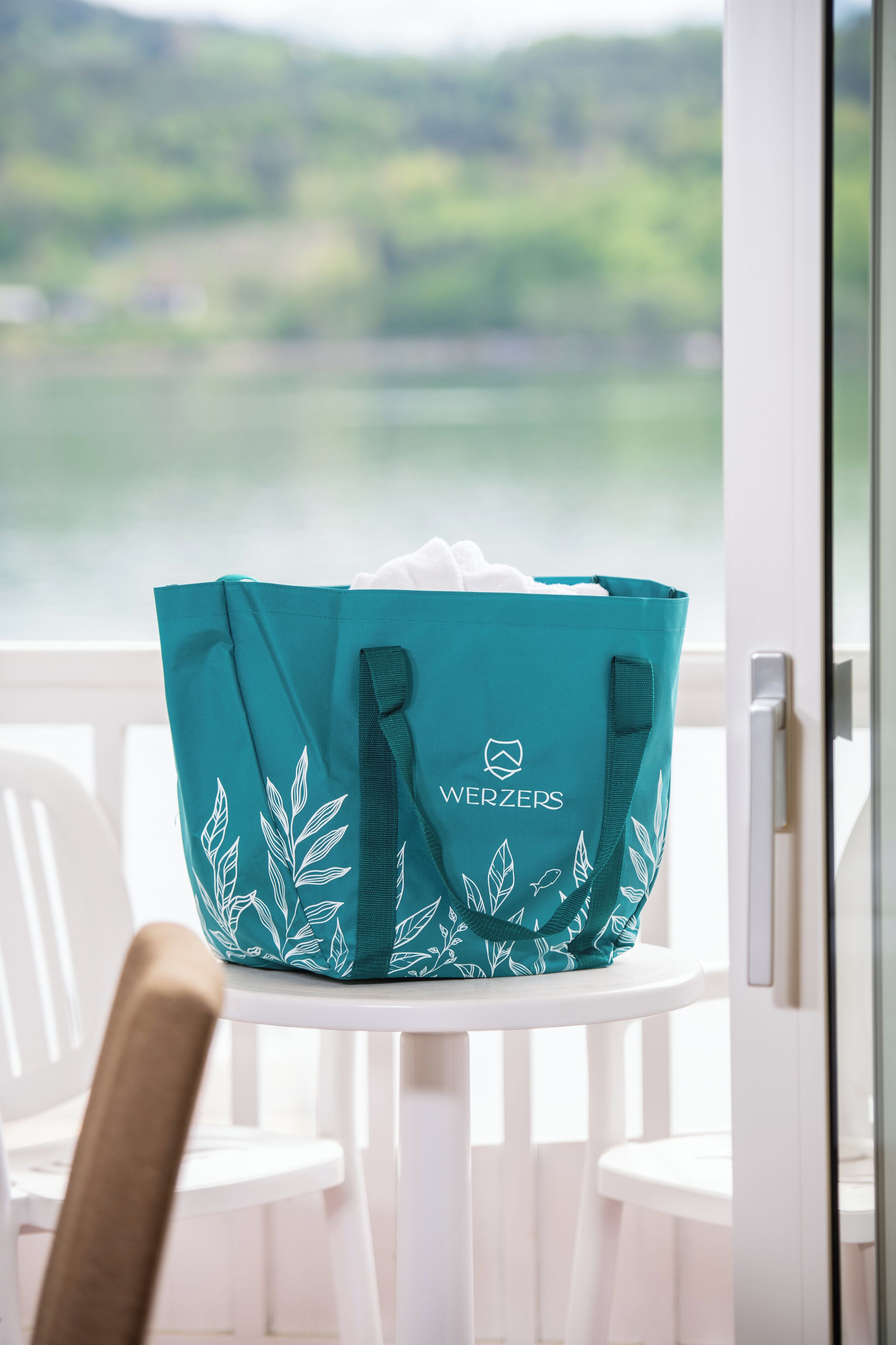 Una bolsa de mano color aguamarina con diseños de hojas y el texto 'WERZERS' sobre una silla blanca junto a una ventana con vistas a un paisaje verde.