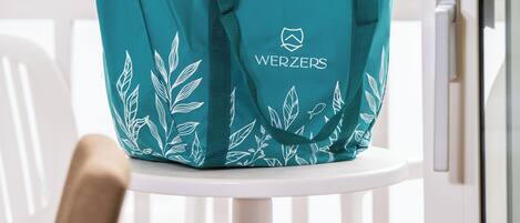 Una bolsa de mano color aguamarina con diseños de hojas y el texto 'WERZERS' sobre una silla blanca junto a una ventana con vistas a un paisaje verde.
