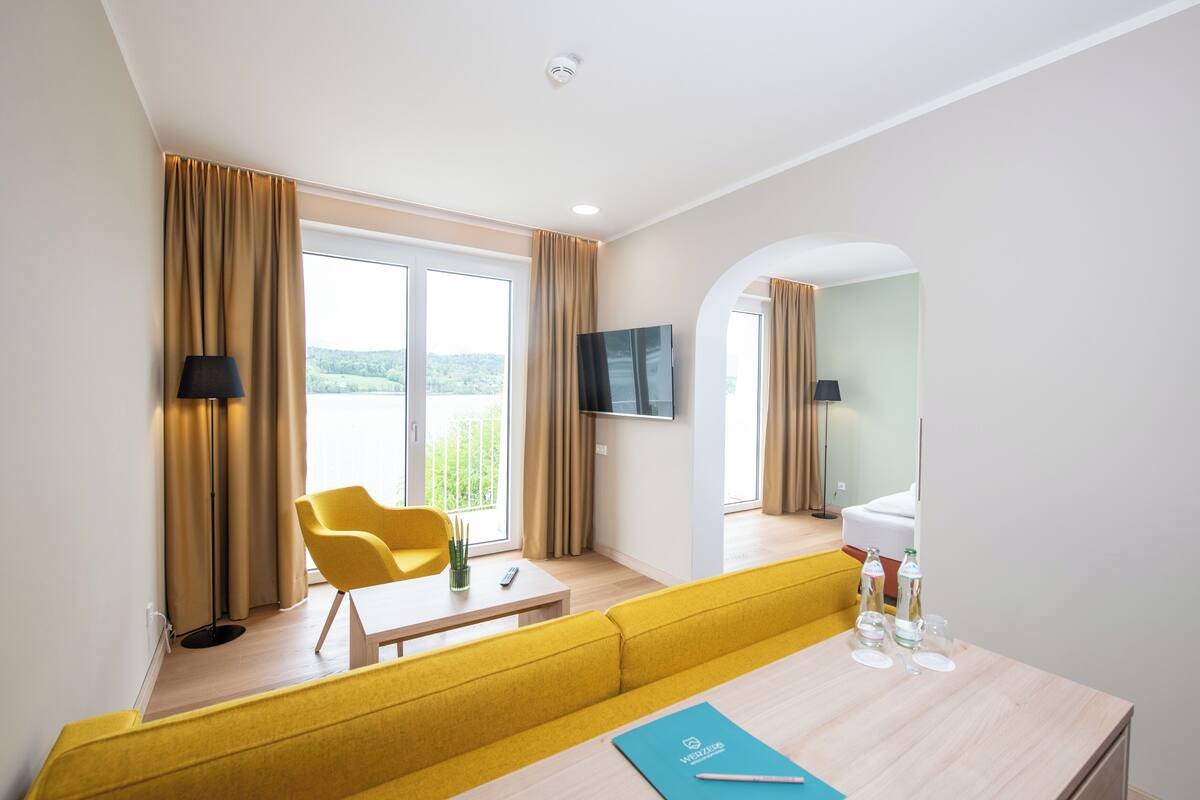 Ein modernes Hotelzimmer mit einem gelben Sofa, einem kleinen Tisch mit einer Fernbedienung, einem Fernseher und Blick ins Freie.