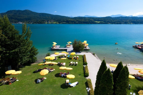 Ein Seehotel mit Liegestühlen, Sonnenschirmen, einem Steg und freiem Blick auf See und Berge.