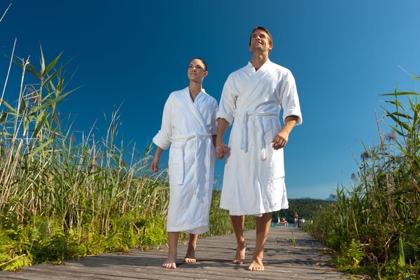 Two people in white robes walking on a wooden boardwalk surrounded by tall grass and greenery. - Werzers Seehotel Wallerwirt (Techelsberg am Worther See)