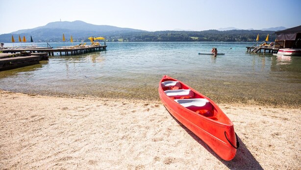 A calm lake with a red canoe on the sand, a dock with yellow umbrellas, and a person in a boat in the distance. - Werzers Seehotel Wallerwirt (Techelsberg am Worther See)