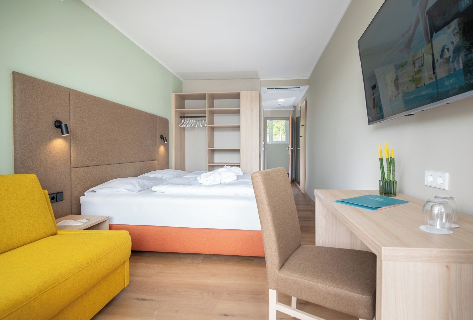 Ein modernes Hotelzimmer mit einem Bett, einem gelben Sofa, einem Schreibtisch mit Stuhl und einem an der Wand befestigten Fernseher.