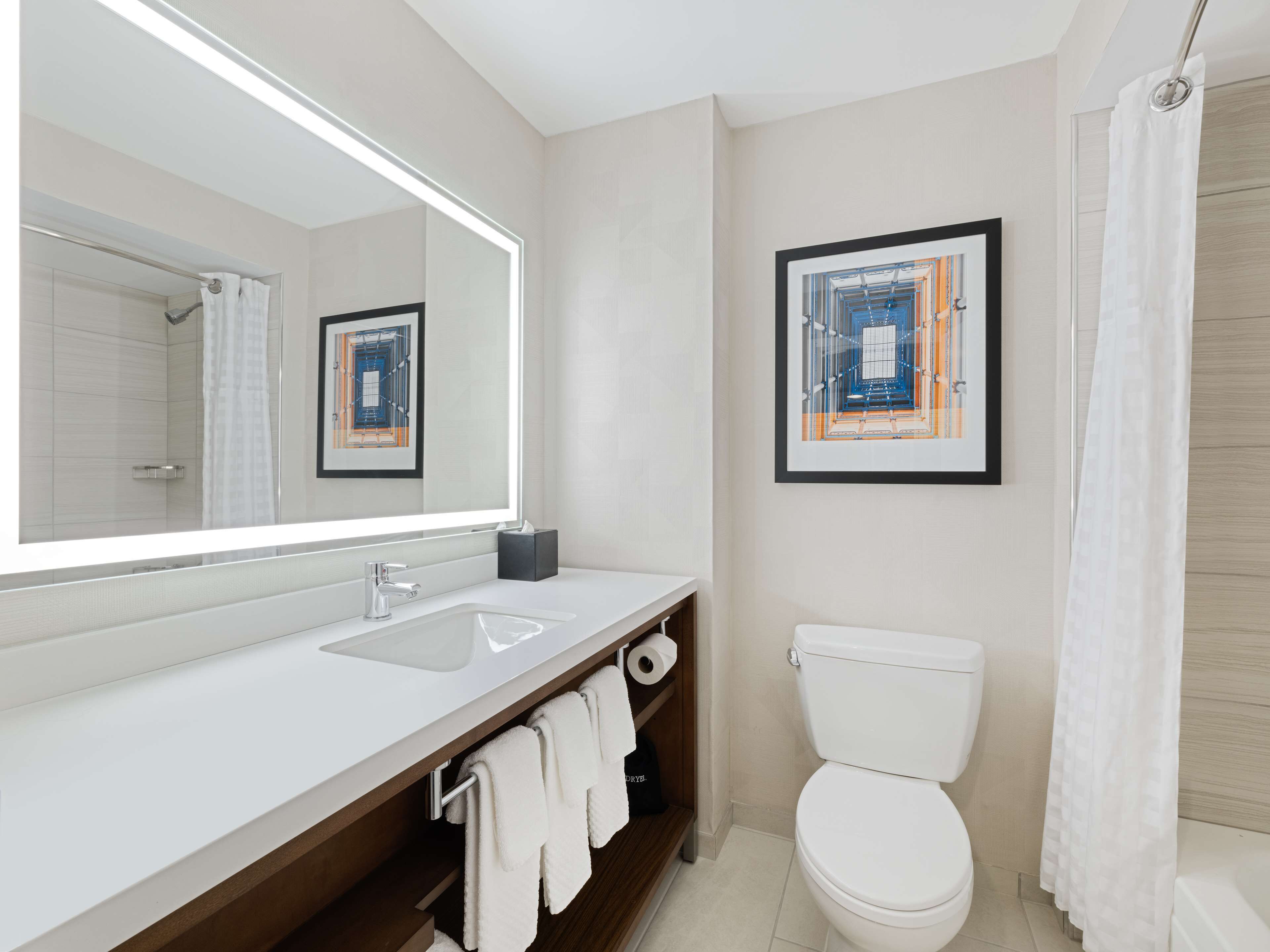 A modern bathroom with a large mirror, white sink, and toilet. There are towels hanging on a rack and a framed picture on the wall.