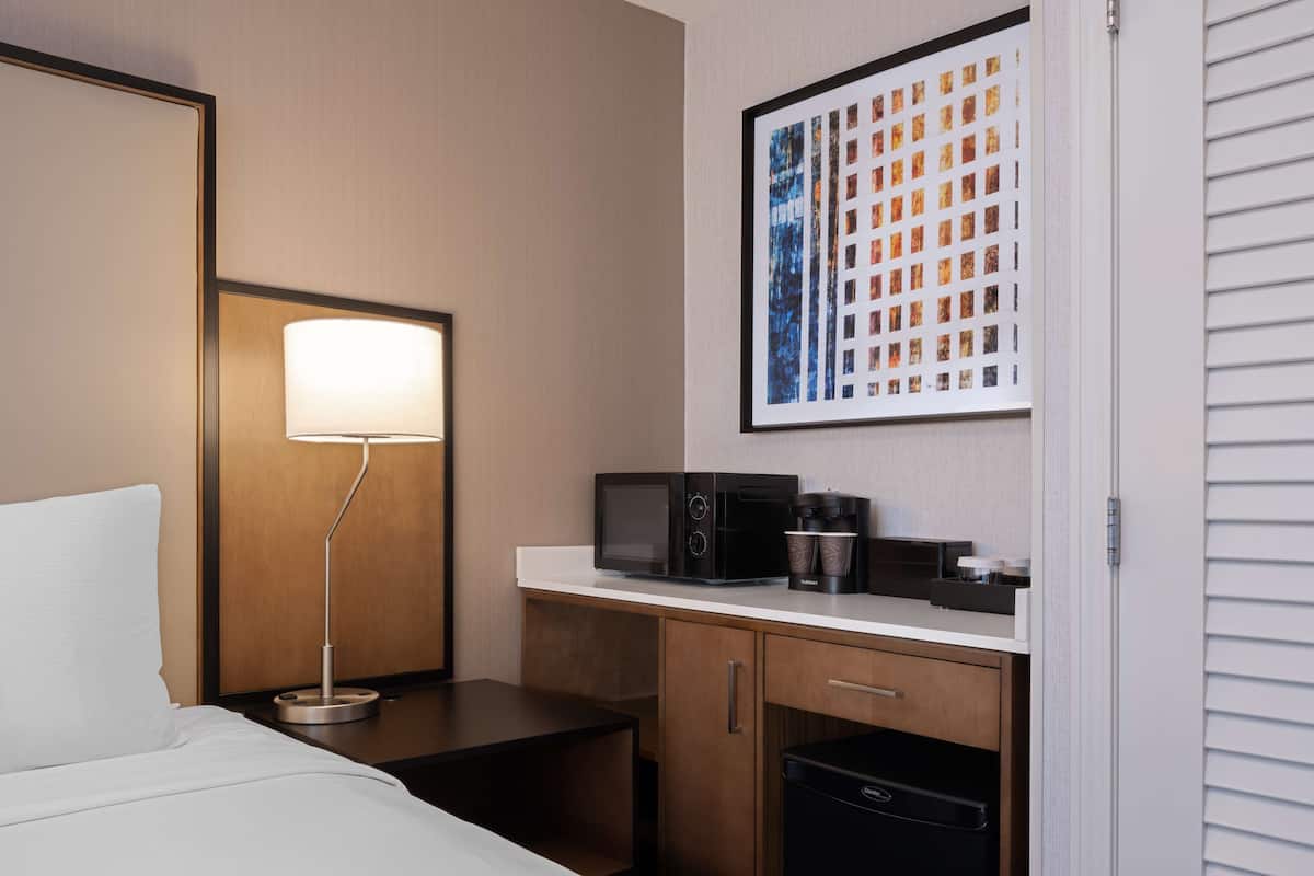 A hotel room with a bed, a desk with a lamp, a coffee maker, a microwave, and a refrigerator.