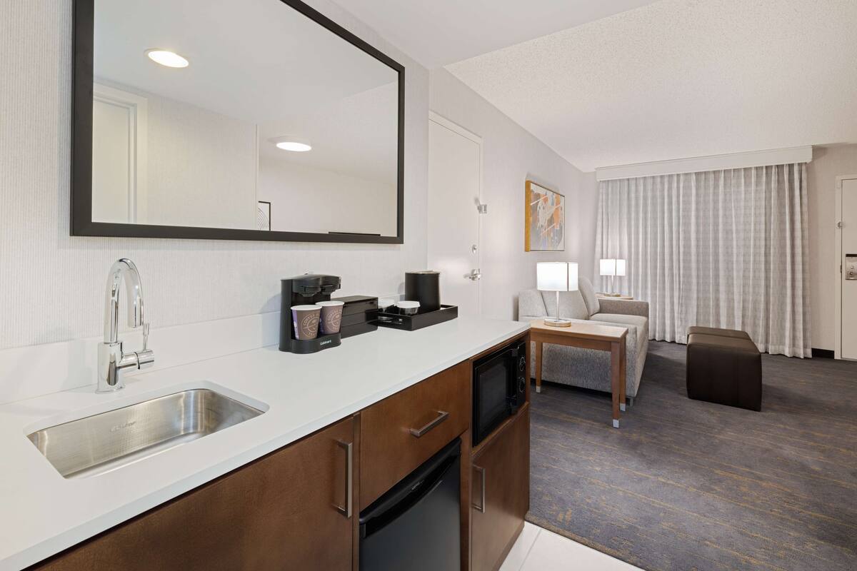 A modern hotel room with a large mirror, a built-in sink, a coffee maker, a microwave, a small table with a lamp, and a seating area with a couch and a chair.