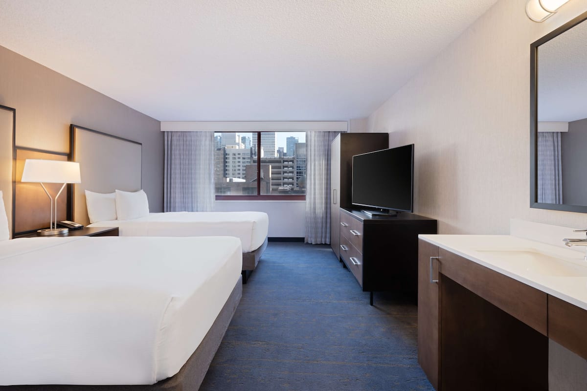 A hotel room with two beds, a TV, a desk, and a large window with a city view.