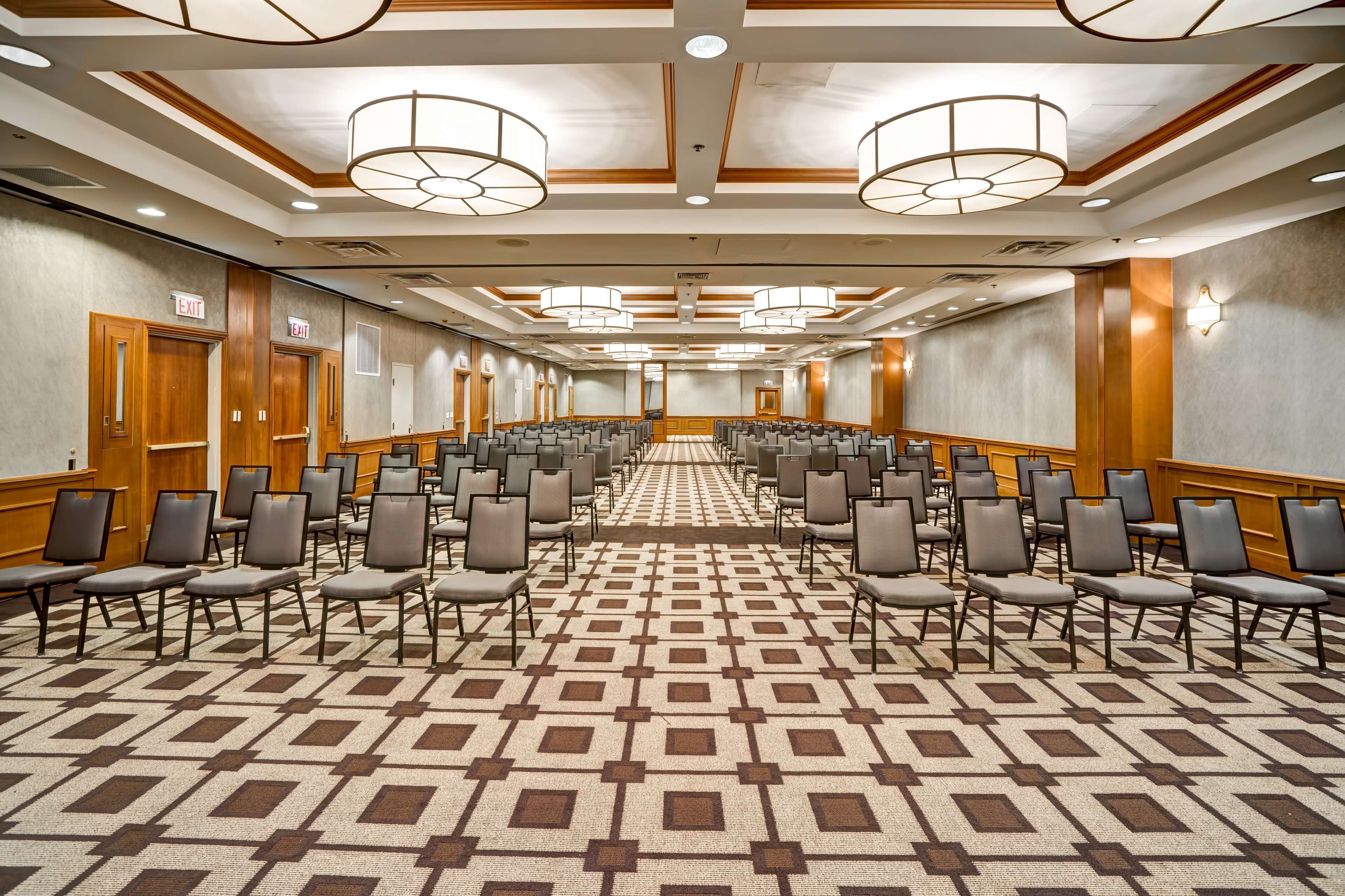 A conference room with rows of chairs, a patterned carpet, wooden paneling, and exit signs.