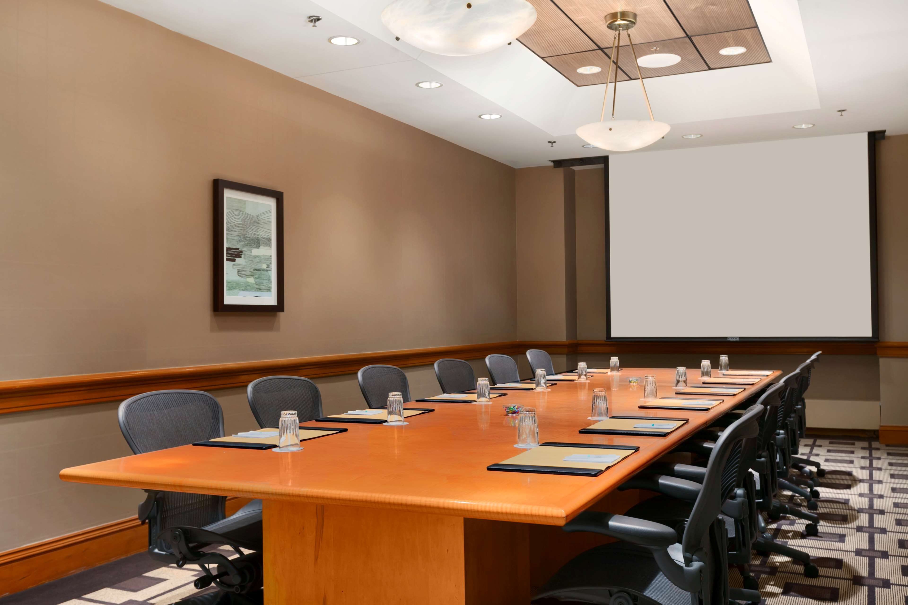 A conference room with a long table, chairs, a projector screen, and a framed picture on the wall.