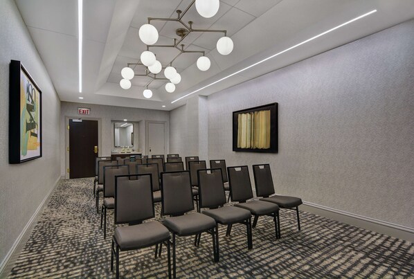 A conference room with rows of chairs, a carpeted floor, a ceiling light fixture, and an exit sign.
