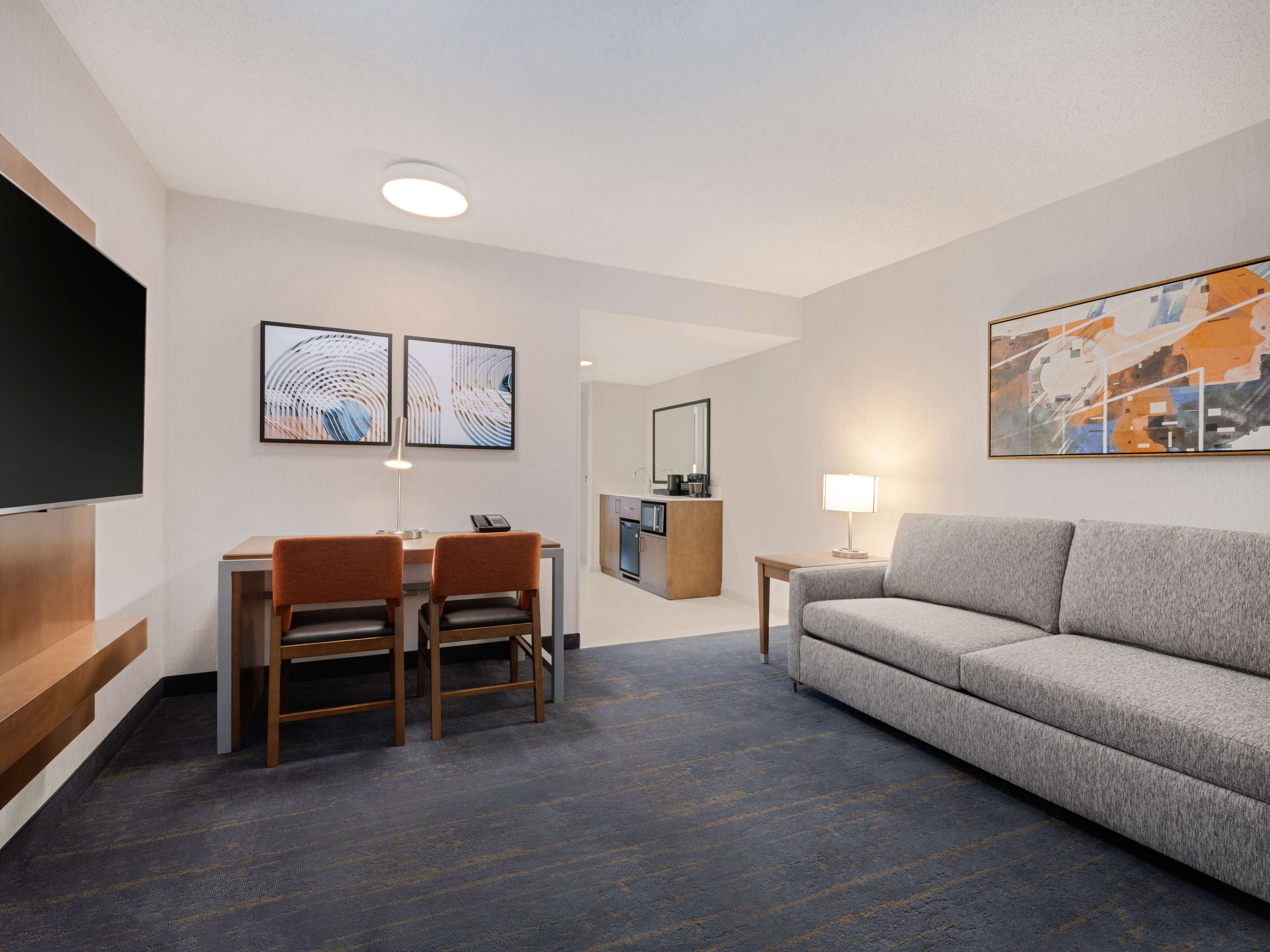 A modern hotel room with a dining area, a sofa, a TV, and artwork on the walls.